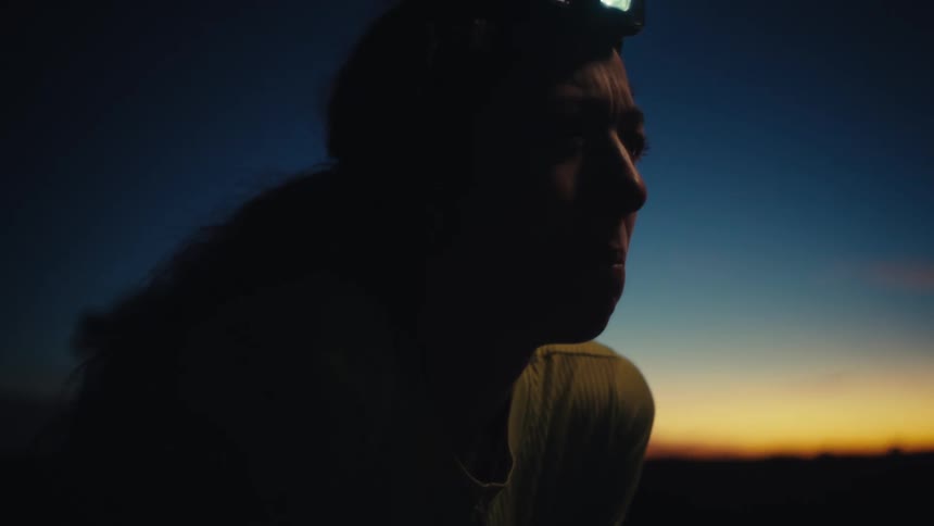 A woman wearing a headlamp and a light shirt stands in a dark field at dusk while looking toward the orange light of a sunset on the horizon