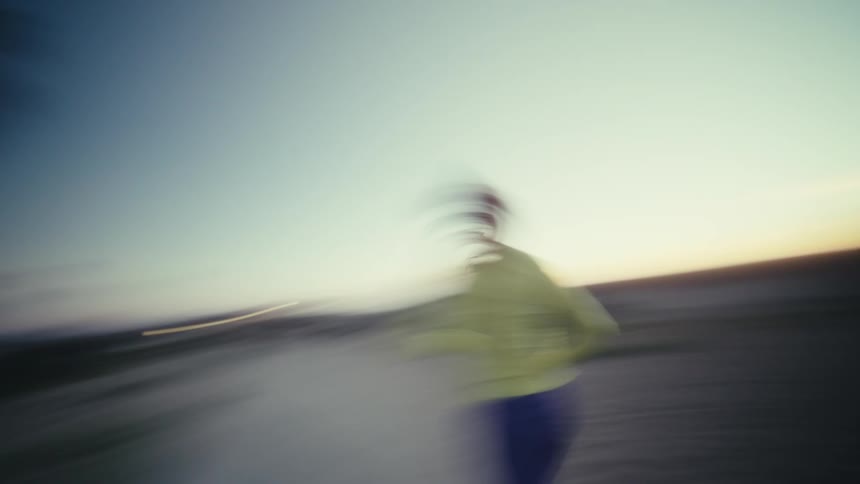 Woman runs at dusk wearing a headlamp in an open area with buildings in the background