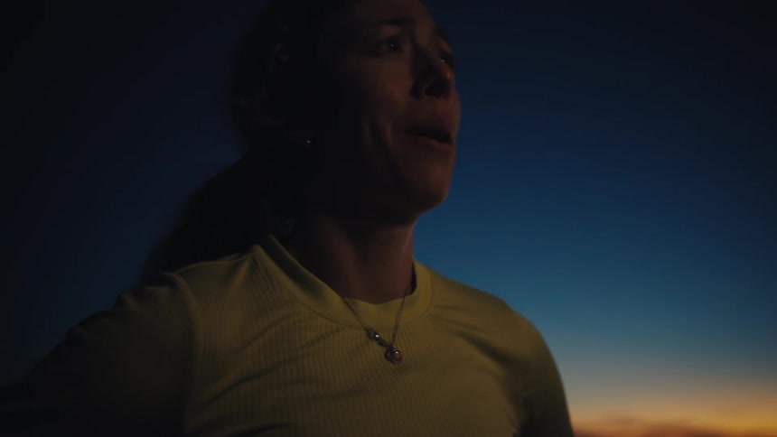 A side view shows a woman wearing a headlamp and a light shirt as she stands in a dark field at dusk while looking toward the orange light of a sunset on the horizon