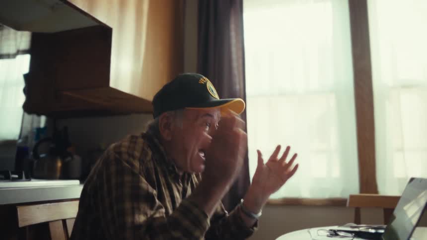 An older man wearing a plaid shirt and a baseball cap cheers with his arms raised while looking at a laptop computer inside a home