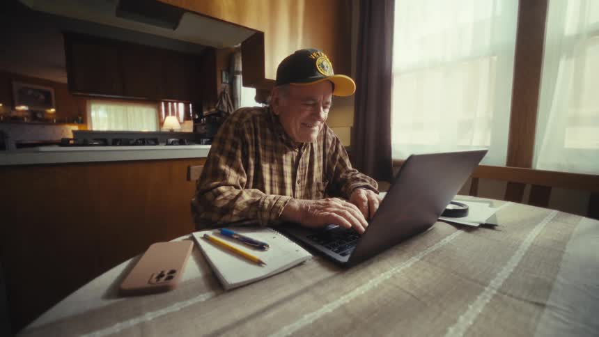 An older man wearing a plaid shirt and a baseball cap smiles while typing on a laptop computer at a table inside a home