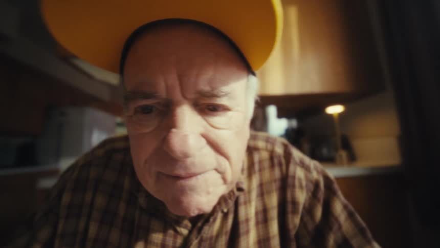 An elderly man wearing a plaid shirt and a veteran hat sits at a kitchen table while typing intently on a black laptop