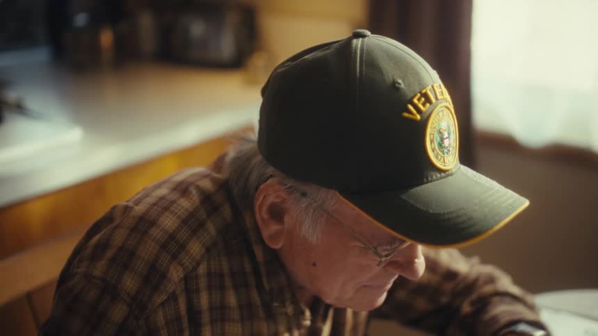 An elderly man wearing a green veteran hat sits at a desk while using a laptop trackpad and writing in a notebook with a blue pen inside a sunlit room
