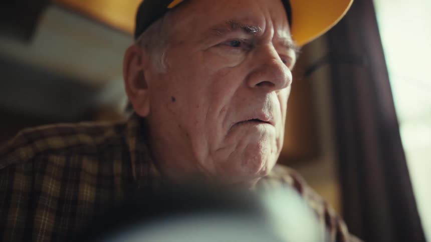 An elderly man wearing a yellow cap and plaid shirt looks closely at a document through a handheld magnifying glass