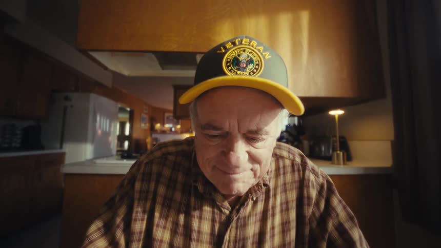 Elderly man seated at a table with a laptop and papers looks out the window in a kitchen lit by natural light