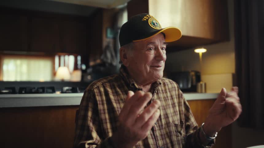 An elderly man sitting at the dining table wearing a baseball hat that says veteran making hand gestures and smiling