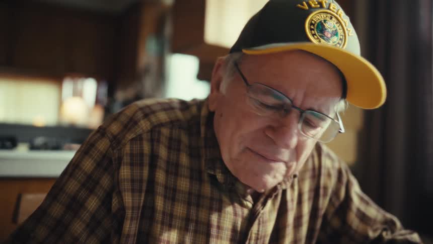Elderly man uses a laptop and writes in a notebook while seated in a kitchen