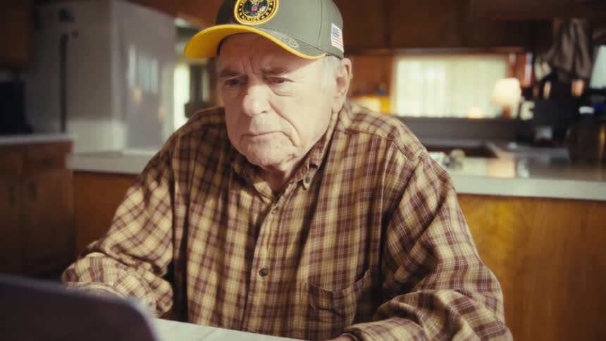 Elderly man wearing a baseball cap and a plaid shirt typing on a laptop computer with a nervous facial expression indoors during day time
