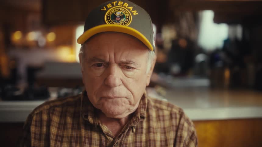 Elderly man frowns as he sits near the kitchen consulting his laptop during the daytime