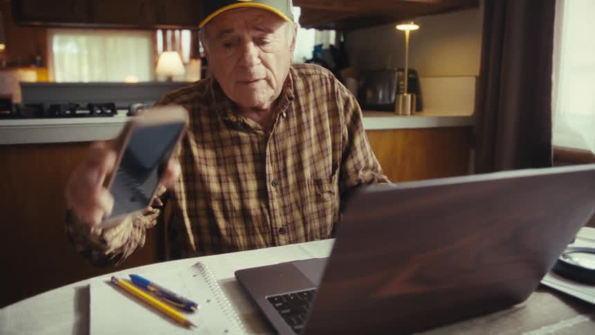 Man sitting at a table in front of a laptop computer as he talks on his phone and then throws his hands in the air