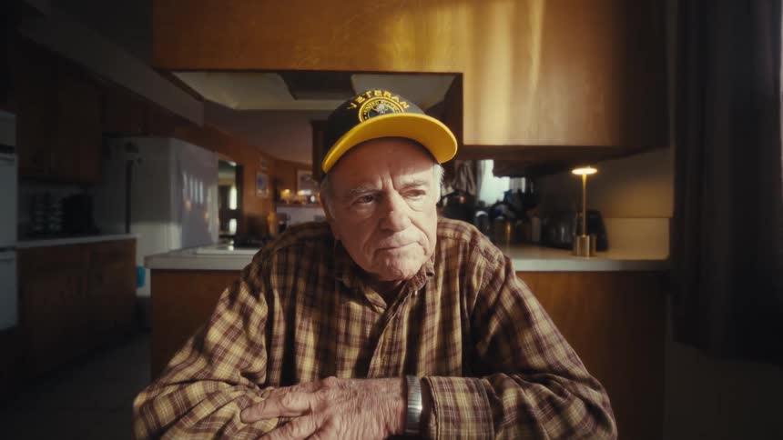 Elderly man closes a laptop while seated at a table in a kitchen lit by natural light