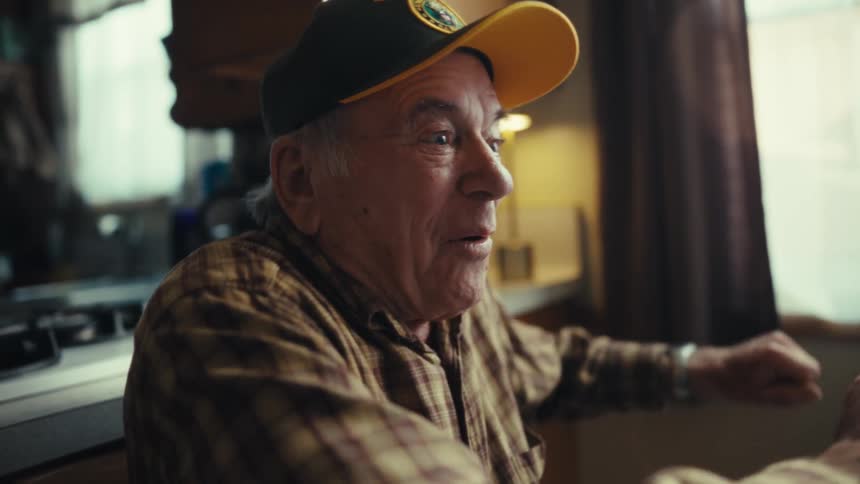 Elderly man seated at a table raises his arms in front of a laptop in a kitchen lit by natural light