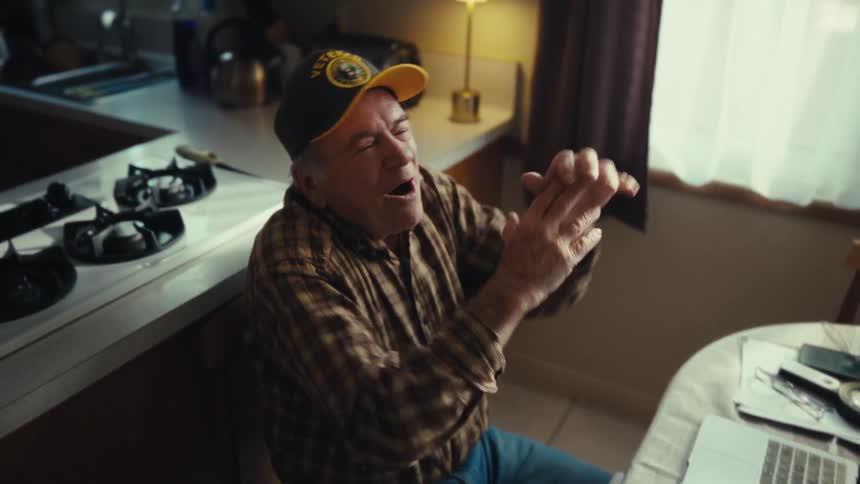 Elderly man seated at a table moves his arms in front of a laptop in a kitchen