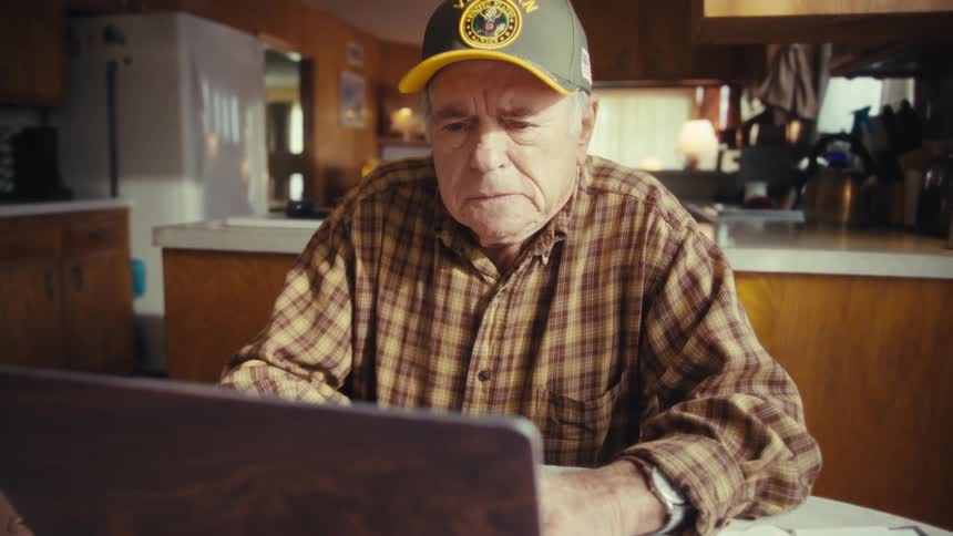 Elderly male veteran frowns while using a computer as he sits at a table near the kitchen