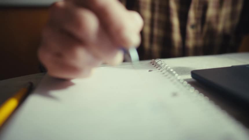 A close up view shows a person using a blue pen to write on a lined notebook next to a laptop computer on a table