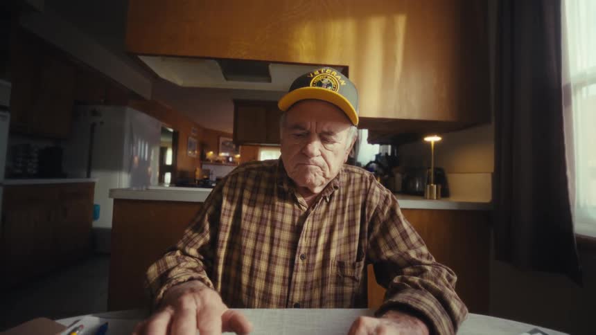 An elderly man wearing a yellow veterans hat sits at a kitchen table while focused on a laptop as the camera zooms in he stops typing and looks off to the side with a contemplative expression