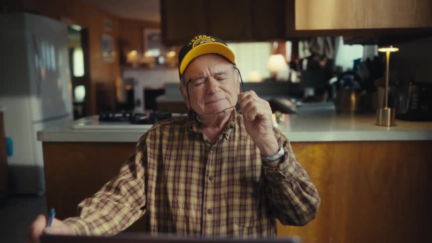 Elderly man wearing a baseball cap and plaid shirt putting on his glasses and reading on a laptop computer indoors during day time