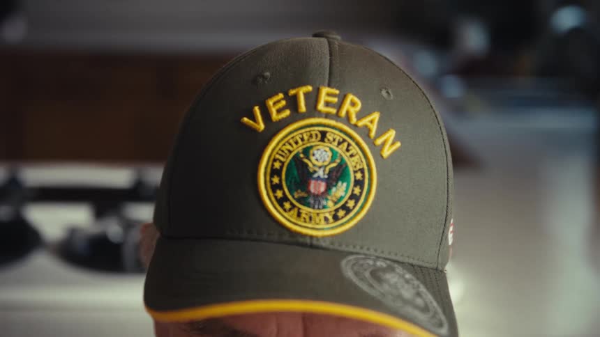 An elderly army veteran wears a green baseball cap with a gold patch as he looks down in a kitchen