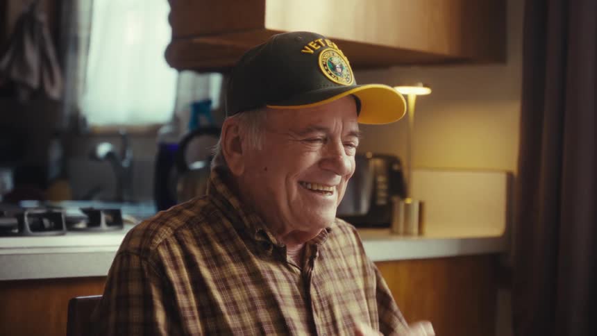 An elderly man in a yellow veterans cap sits at a kitchen table and looks at a laptop screen as he watches his expression shifts from a gentle smile to a hearty joyful laugh