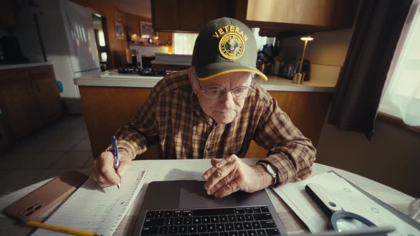An elderly man sitting at the dining table at midday wearing glasses and a hat that says veteran writing on a note with a pen while looking at a laptop screen