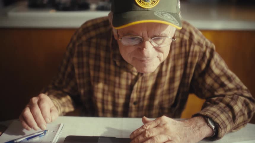 Elderly man with glasses and yellow cap focused on laptop at table