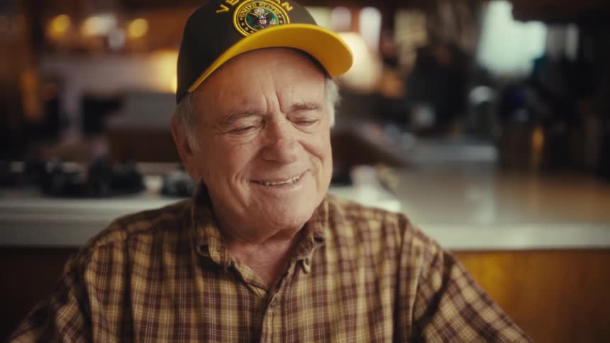 An older man wearing a plaid shirt and a baseball cap smiles and laughs while looking around inside a kitchen