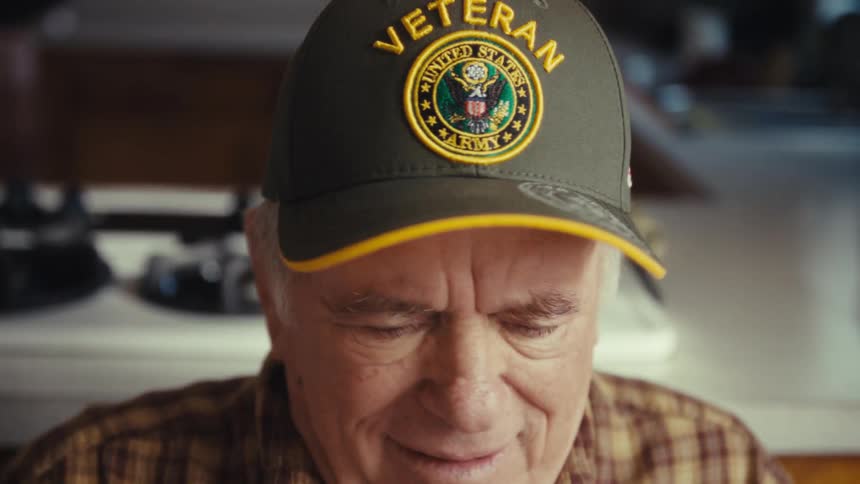 An older man wearing a plaid shirt and a baseball cap looks down with a slight smile while sitting at a table inside a home