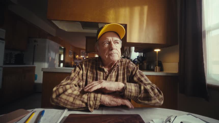 An elderly man wearing a veteran hat sits at a kitchen table and closes a dark wooden laptophe then rests his arms on the table and looks thoughtfully out of a nearby window