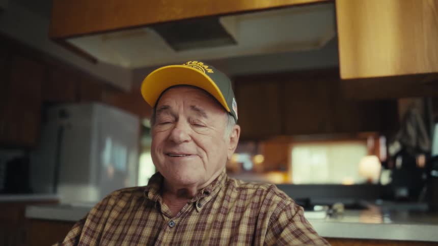 Elderly man removes his glasses while seated in a kitchen