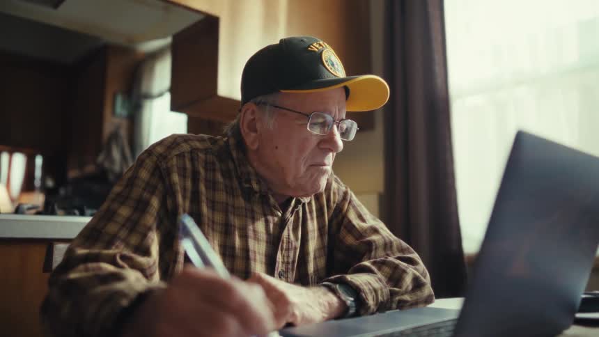 Elderly man writing in notebook in front of laptop in home kitchen