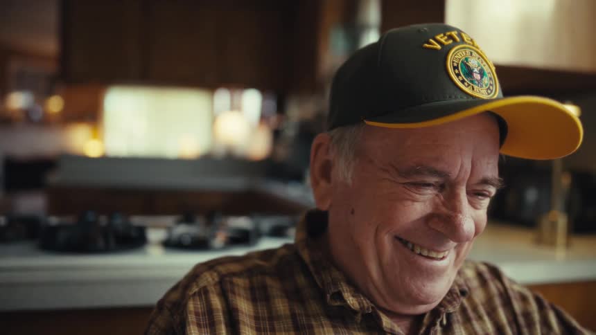 A smiling elderly veteran wearing a yellow and black cap laughs while removing his glasses