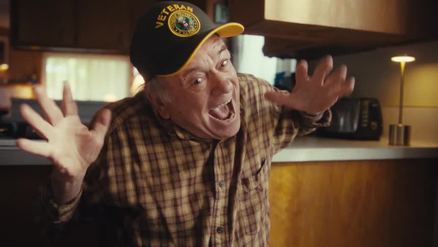 An elderly man wearing a veteran hat and plaid shirt laughs joyfully while raising his arms in a celebratory gesture inside a kitchen