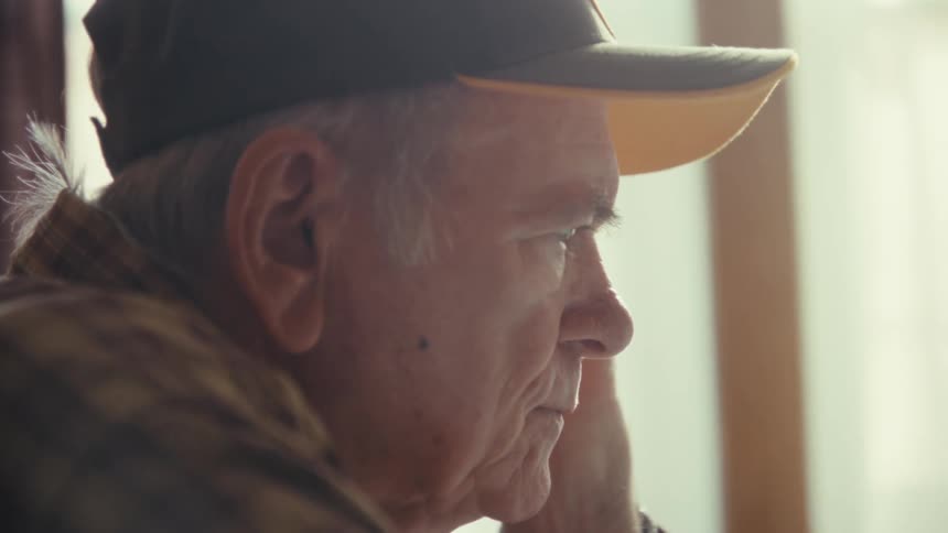 An elderly man wearing a plaid shirt and a baseball cap sits indoors and looks out a window
