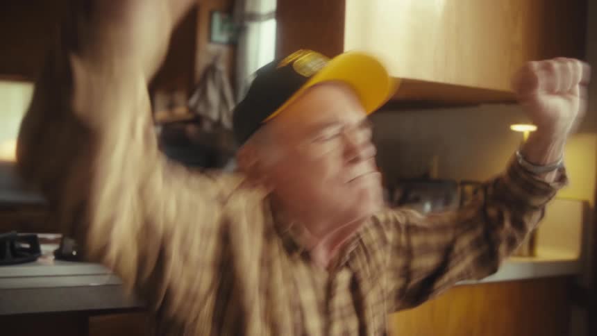 A joyful elderly man wearing a yellow veteran baseball cap and a plaid shirt dances enthusiastically with his arms raised while standing in a brightly lit domestic kitchen near a stovetop and toaster