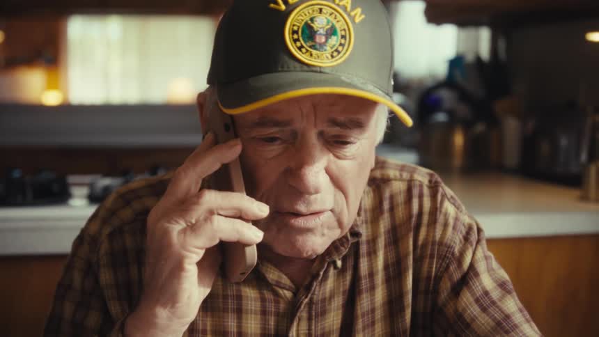 An older man wearing a plaid shirt and a baseball cap talks on the phone while looking around inside a kitchen
