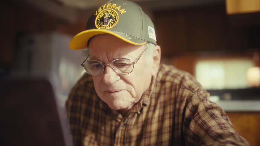 Elderly man wearing a veteran cap and glasses uses a computer indoors