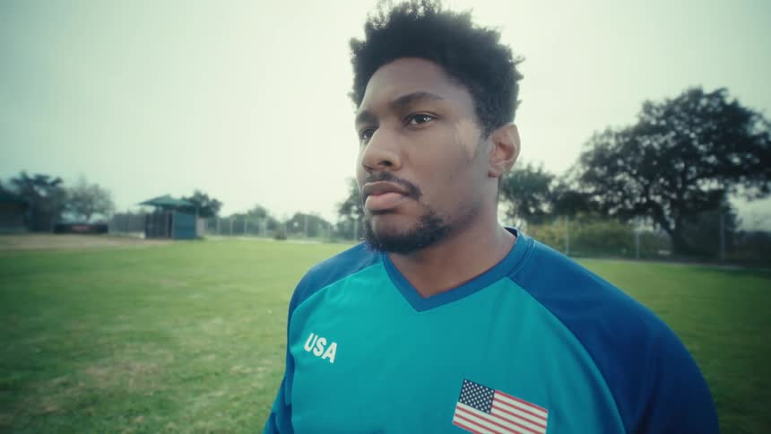 A closeup shot of a young man with an athletic build standing on an outdoor sports field he is wearing a blue athletic jersey with usa and the american flag on it looking offcamera with a serious and focused expression