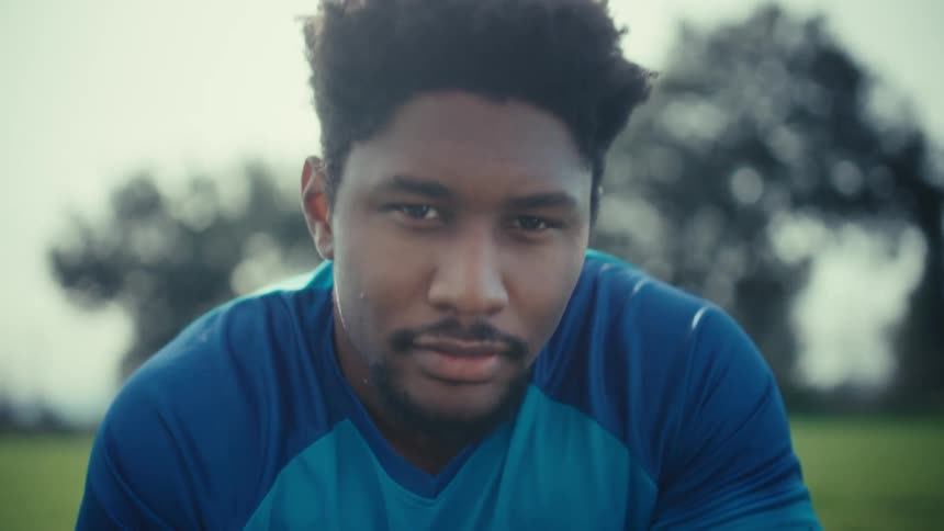 A young man wearing a blue usa soccer jersey crouches on a grass field while holding a ball and looking directly into the camera with a serious expression