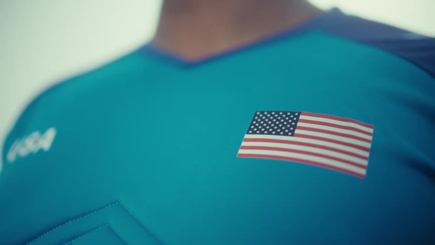 A man wears a blue athletic jersey featuring a small united states flag patch on the chest and usa text on the side