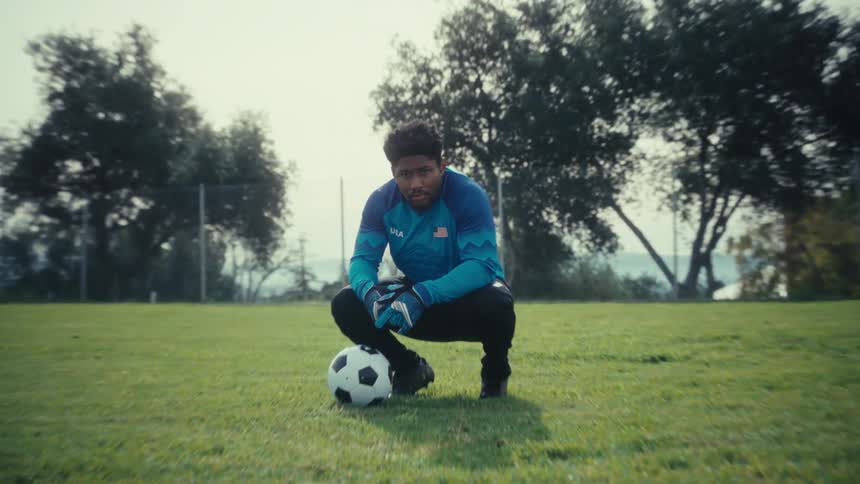 A young man wearing a blue jersey and goalie gloves squats on a green field next to a soccer ball