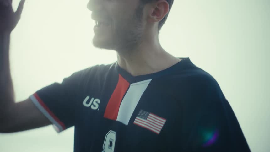 A young man wearing a dark jersey with usa and an american flag on it smiles and talks while standing on a grassy field