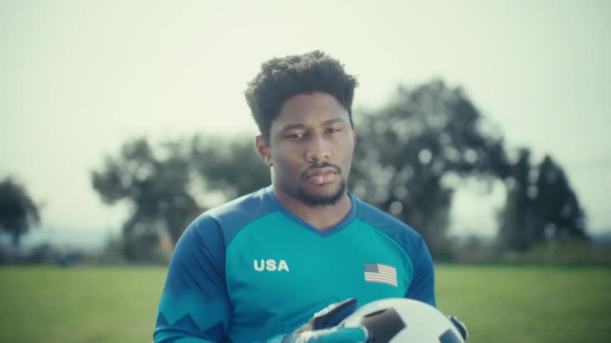A soccer goalkeeper in a blue usa jersey stands on a grassy field holding a ball he looks directly into the camera with a focused expression as he slowly lowers the ball from his face