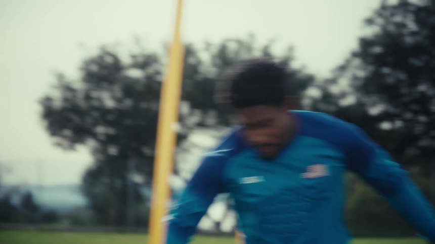 A soccer player in a blue usa jersey performs agility drills by weaving through yellow poles on a grass fieldthe camera follows his movements closely as he transitions from footwork exercises to dribbling a soccer ball across the turf