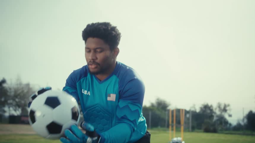 A soccer goalkeeper in a blue jersey and gloves on a training field catching a soccer ball and then kicking it with his feet at midday as part of his practice