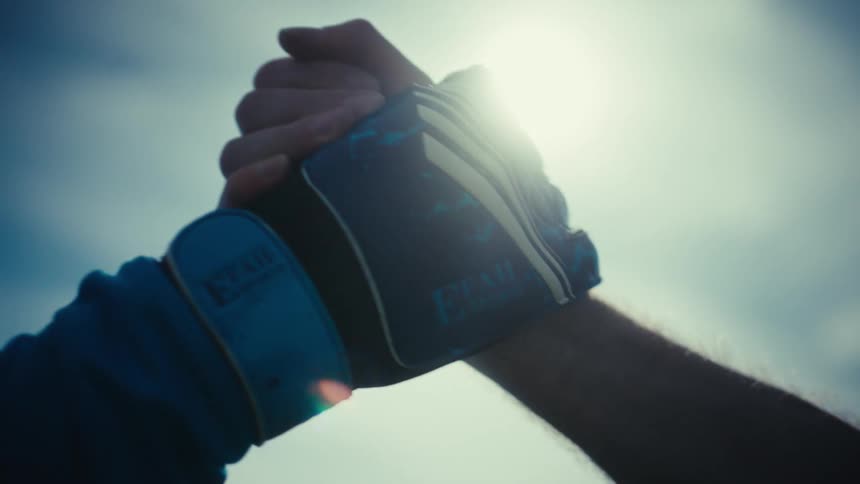 A close up view shows two people as they grip each other by the hands in a show of support against a bright sky