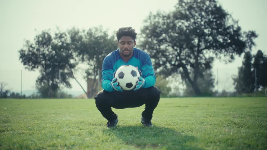Young adult black man wearing a goalkeeper uniform throwing and picking up a soccer ball on a field outdoors during day time