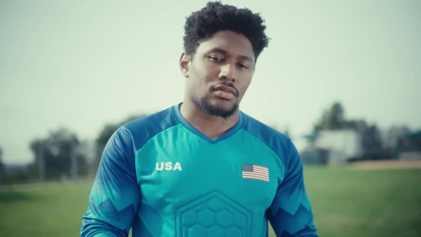 A young man wearing a blue jersey with usa and an american flag on it looks down while holding a soccer ball on a green field