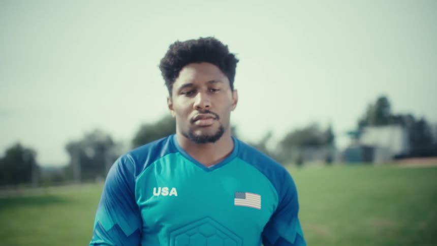 A soccer goalie in a blue usa jersey stands on a grass field holding a soccer ball he looks directly into the camera with a focused expression as the shot zooms out to show his full upper body