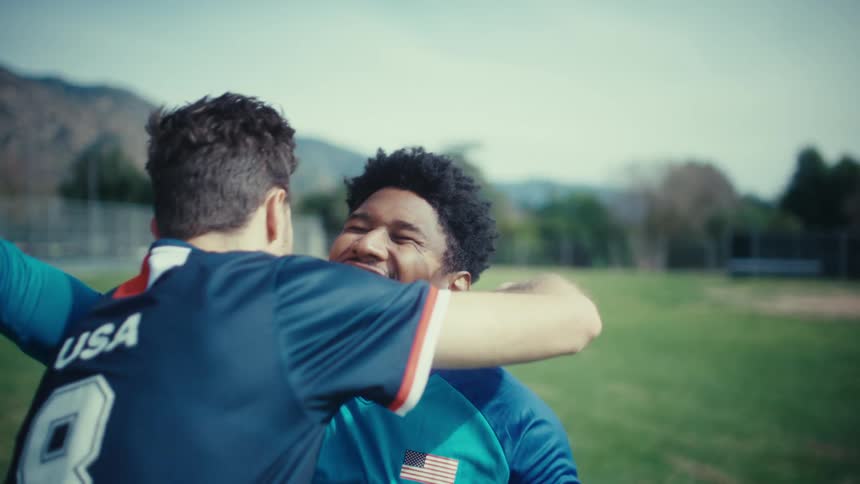 Two soccer players embrace in a hug on a green field as one athlete wearing a blue jersey and goalie gloves smiles while the other player in a dark usa jersey holds him