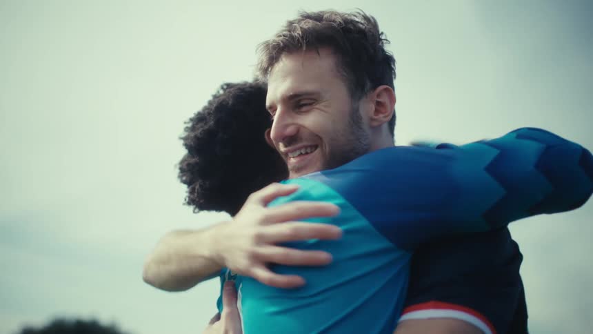 A young man wearing a blue jersey and goalie gloves hugs another soccer player on a green field